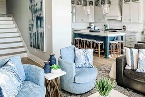 Un salon moderne avec un canapé bleu, une table basse agrémentée d’objets décoratifs et un coin cuisine doté de meubles blancs et de tabourets de bar.
