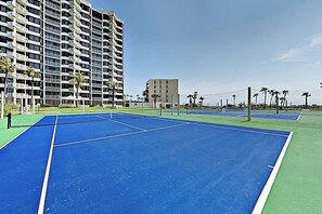Ein Tennisplatz mit Netz, umgeben von einer Mauer und Palmen, mit einem Hochhaus im Hintergrund.
