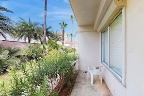 A small outdoor area with a white chair, a window with closed blinds, and various plants including palm trees. - Dolphin Condominiums #20 (South Padre Island)