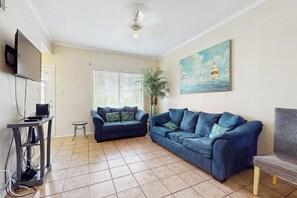 A living room with a blue sofa, a television mounted on the wall, a painting of a lighthouse, and a tiled floor. - Dolphin Condominiums #20 (South Padre Island)