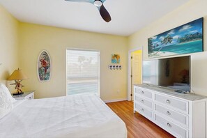 A bedroom with a bed, a ceiling fan, a TV, a dresser, and a painting on the wall. - Hibiscus Hideaway Beach House (South Padre Island)