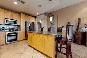A kitchen with yellow cabinets, stainless steel appliances, a granite countertop, and a bar stool. - Sapphire Condominiums #603 (South Padre Island)