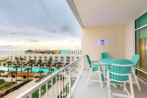 A balcony with a view of a resort pool and buildings, a table with chairs, and a railing. - Sapphire Condominiums #603 (South Padre Island)
