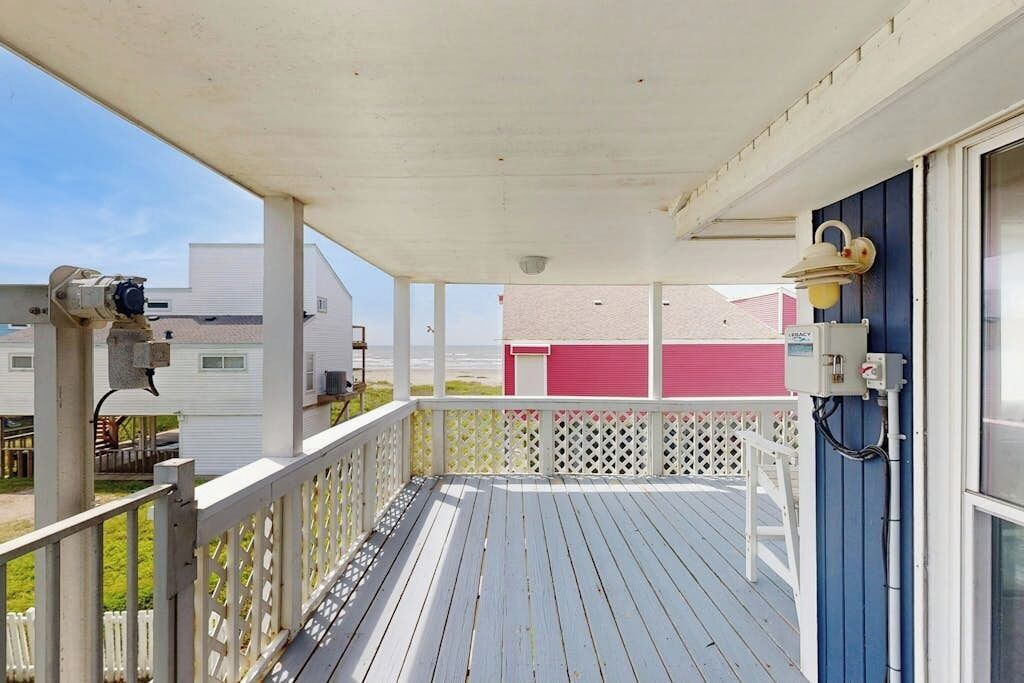 Una terraza techada con barandilla blanca, un muro azul y un edificio rojo al fondo.