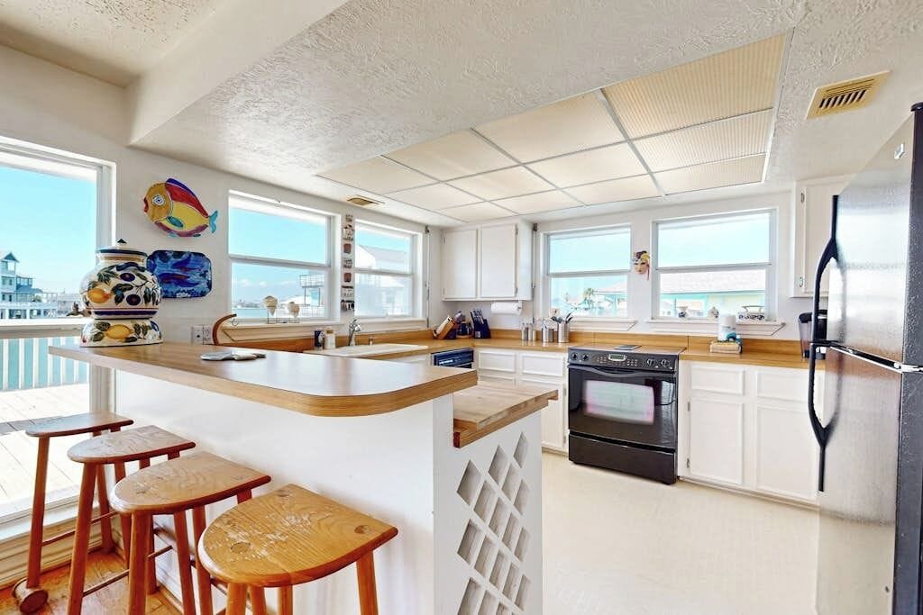 A modern kitchen with a central island, bar stools, a refrigerator, and large windows.