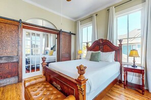 A bedroom with a large bed, a wooden bench, a nightstand, and a view of the outside. - Blue Sky Getaway - Private Pool & Historic Charm (Galveston)