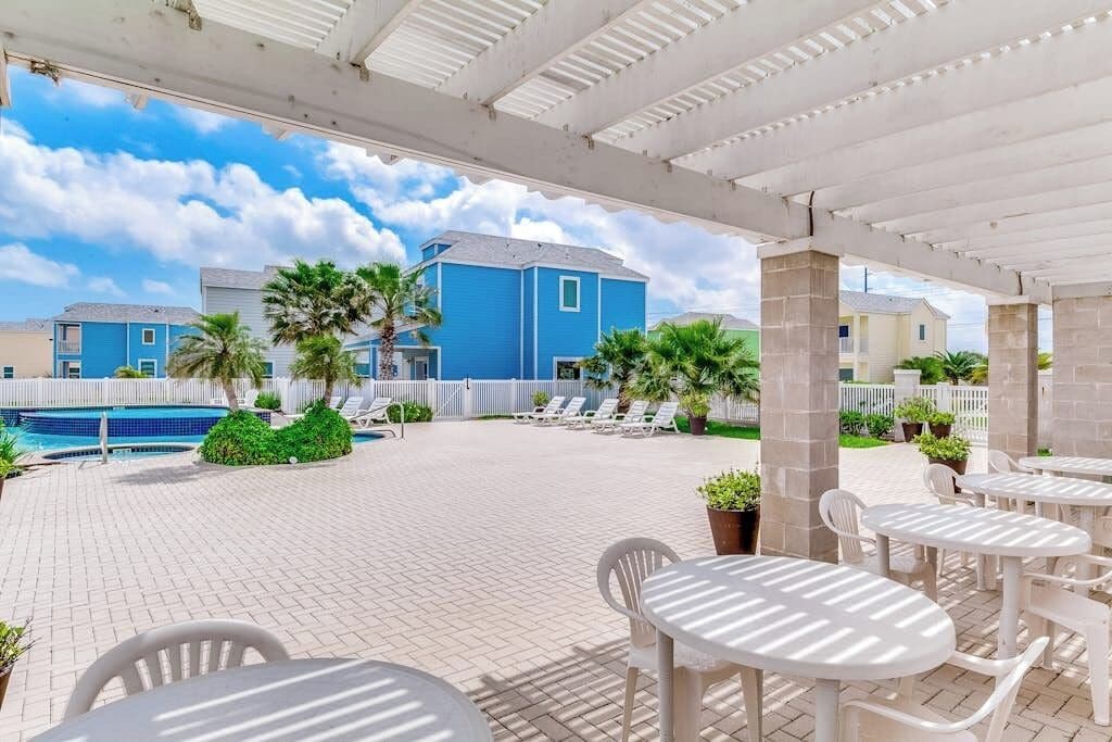 Un espacio al aire libre techado con sillones de descanso y mesas, una piscina y un edificio azul al fondo.