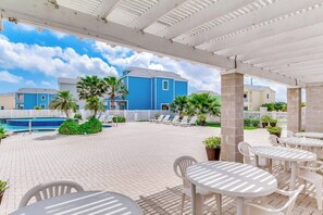 Uma área externa coberta com cadeiras de estar e mesas, piscina e um edifício azul ao fundo.