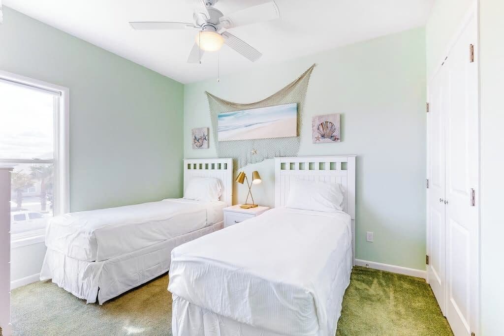 A room with two beds, a ceiling fan, a window, and artwork on the wall.