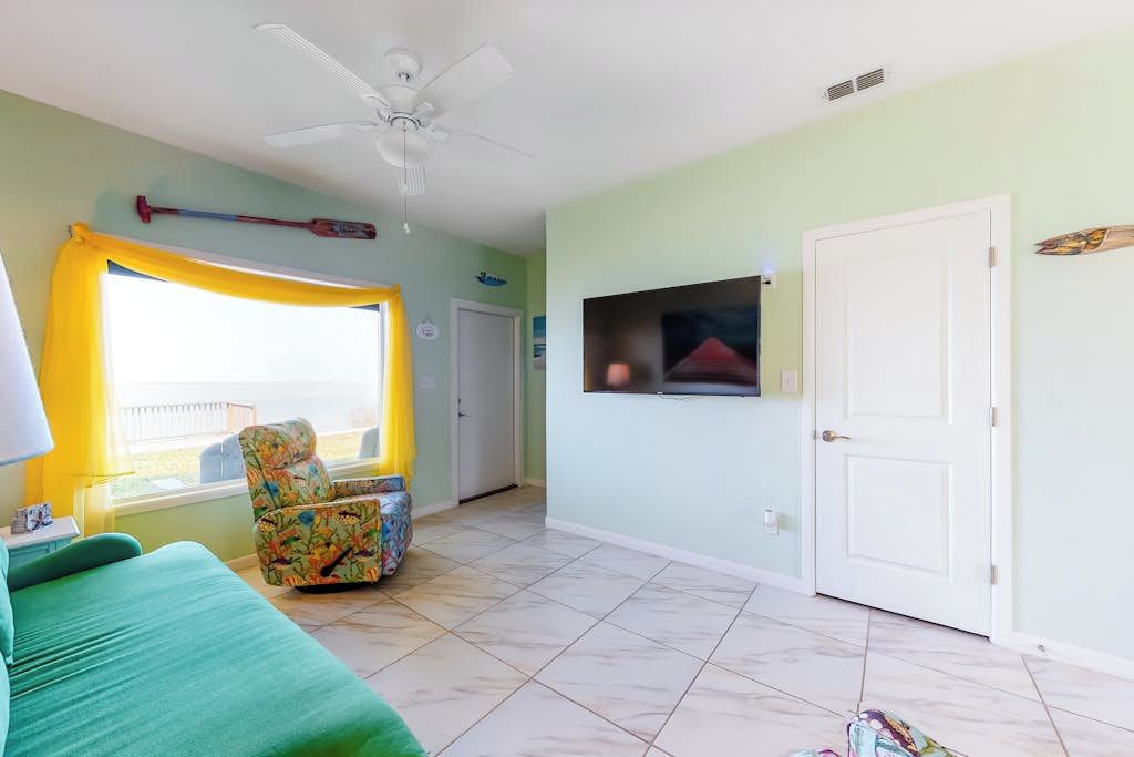 A room with a green sofa, a window with yellow curtains, a ceiling fan, and a television on the wall.