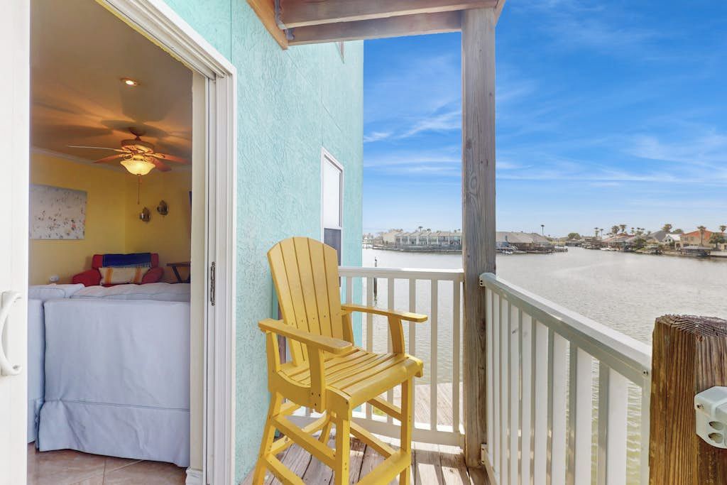 Ein Balkon mit Blick auf ein Gewässer, ein Holzstuhl und ein Zimmer mit Bett und Deckenventilator.