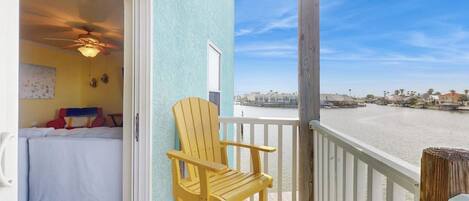 A balcony with a view of a body of water, a wooden chair, and a room with a bed and a ceiling fan.