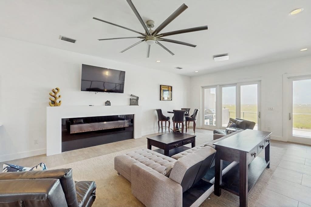 A modern living room with a ceiling fan, a flat-screen TV, a fireplace, and a dining area with chairs.