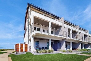 A modern multi-story building with balconies, a covered outdoor seating area, and a well-maintained lawn. - Padre Villa 104 - Gulf & Lake Views, Sleeps 14 (Corpus Christi)