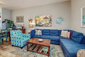 Ein Wohnzimmer mit blauem Sofa, gemustertem Sessel, Couchtisch aus Holz und einem tropisch gestalteten Gemälde.