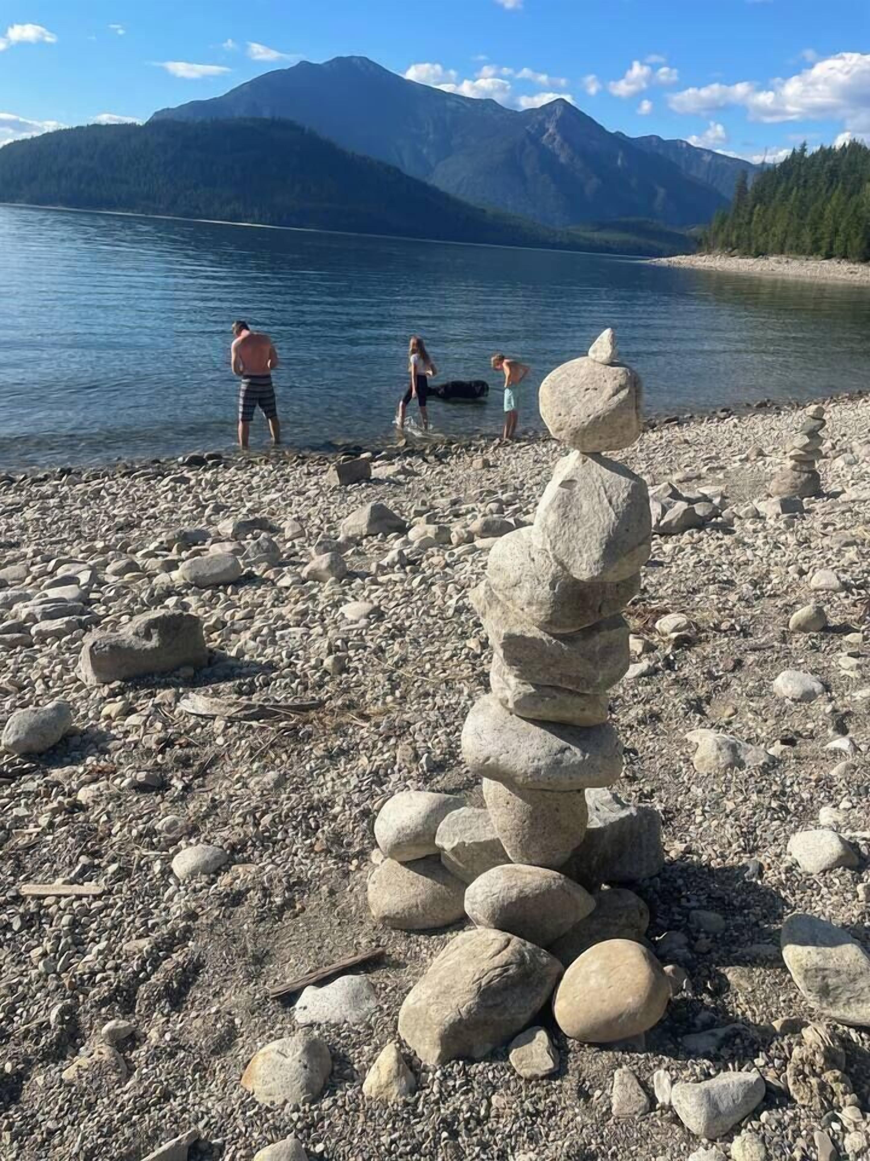 A stack of stones on a pebbly shore with a lake and mountains in the background.