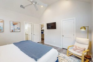Um quarto com cama, uma cadeira, ventilador de teto e televisão na parede.