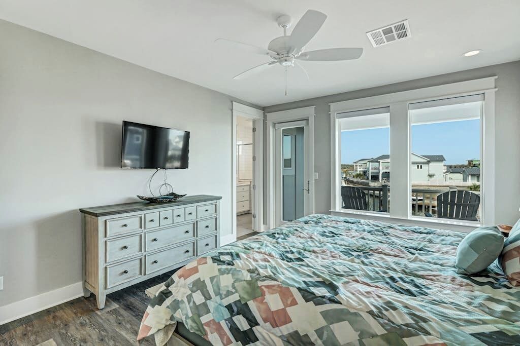 A bedroom with a bed, a ceiling fan, a TV, a dresser, and large windows with a view of houses.