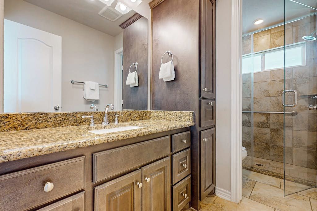 Un baño con encimera de granito, muebles de madera oscura, cabina de ducha de vidrio y una puerta blanca.