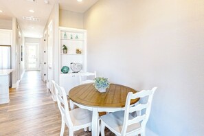 A dining area with a round wooden table and white chairs, a white cabinet with decorative items, and a hallway leading to another room.