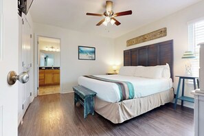 A bedroom with a bed, a ceiling fan, a bench, a bedside table, a nightstand, and a view into the kitchen. - South Pearl Sailfish (Rockport)
