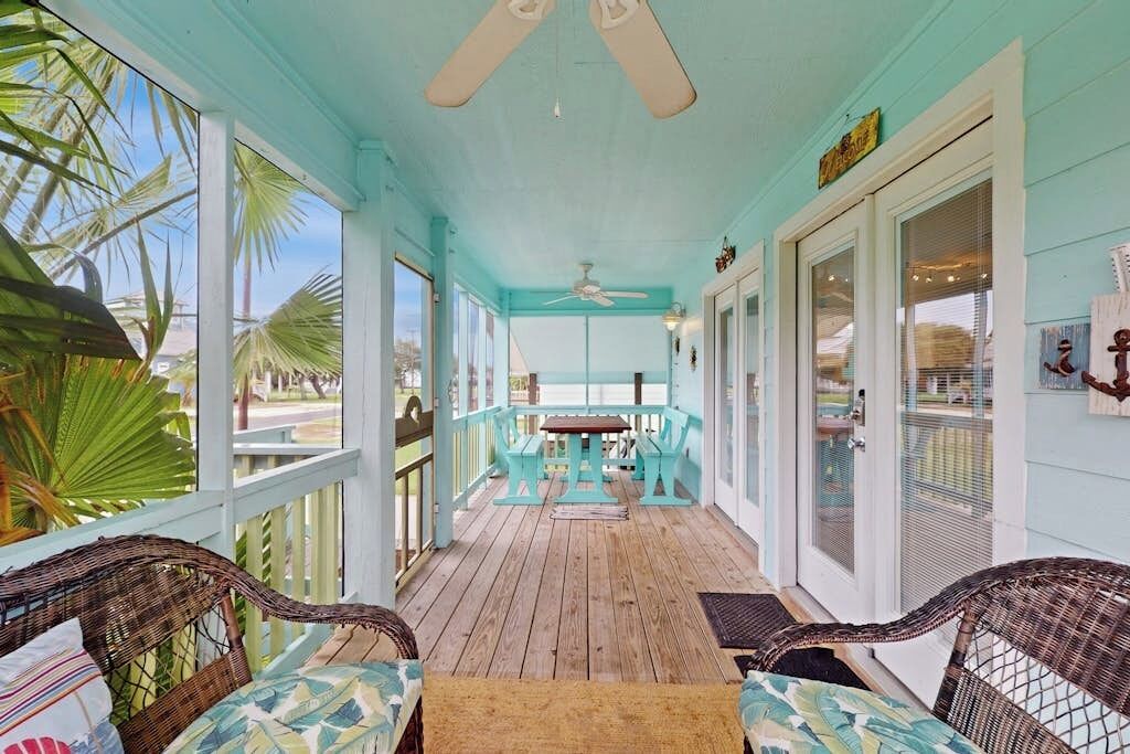 A covered porch with a wooden floor, two wicker chairs, a table, and a ceiling fan.