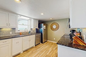 A kitchen with white cabinets, a stainless steel refrigerator, and a black countertop. - Lost Colony Make A Splash (Port Aransas)