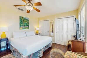 A hotel room with a bed, a ceiling fan, a painting, two doors, a television, and a wicker chair. - Lost Colony Make A Splash (Port Aransas)