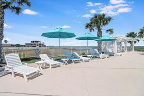A beachside lounge area with white lounge chairs and turquoise umbrellas, a white pergola, and a view of the ocean.