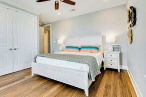 Quarto com cama branca, piso de madeira, ventilador de teto e mesa de cabeceira com abajur.