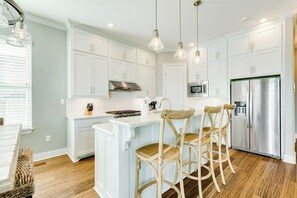 A kitchen with white cabinets, a stainless steel refrigerator, and a bar island with chairs. - Seaside Paradise Retreat (Galveston)