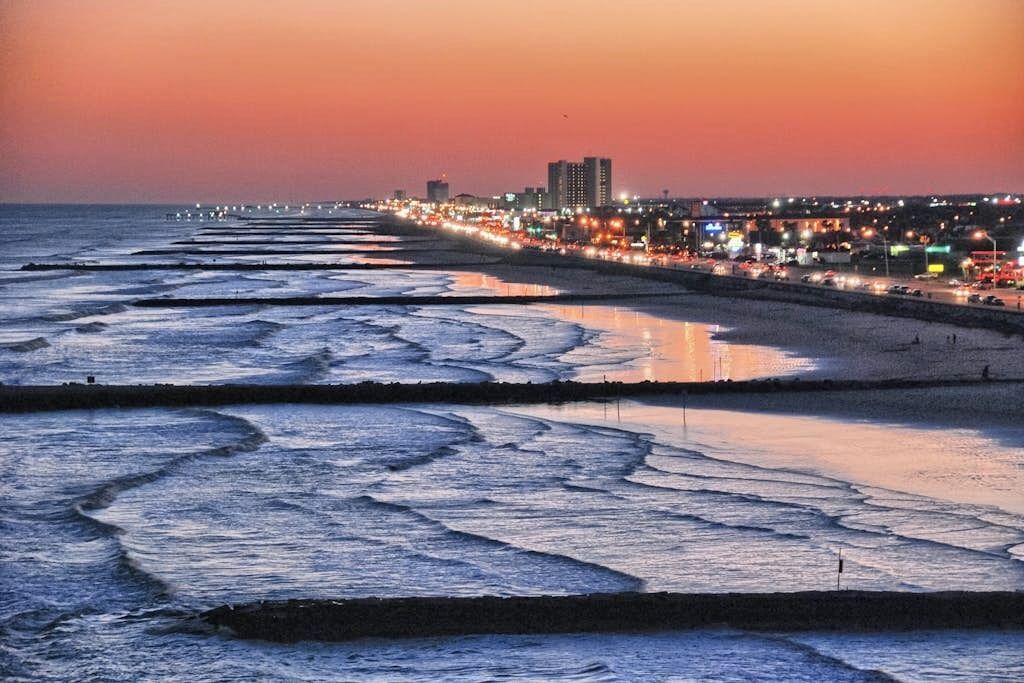 A coastal cityscape at dusk with a beach featuring calm waves and a city skyline in the background.