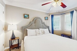 A bedroom with a bed, a nightstand, a lamp, a ceiling fan, and two windows with curtains. - Seascape Gem - Ocean-View Balcony, Pool & Hot Tub (Galveston)