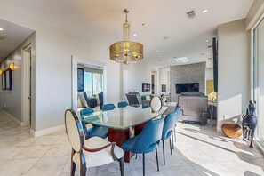 A modern dining area with a glass table and blue chairs, a chandelier, and a kitchen in the background. - Diamond Beach Crown Jewel - #714 (Galveston)