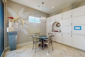 A dining area with a glass table and metal chairs, a decorative wall with a bird motif, and white cabinets with a blue sign. - Diamond Beach Crown Jewel - #714 (Galveston)
