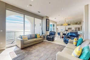A spacious living area with a large window, a sofa, a chair, and a dining area with a chandelier. - Diamond Beach Crown Jewel - #714 (Galveston)