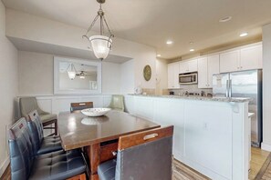 A modern kitchen with white cabinetry, a central island, and a dining area with a wooden table and leather chairs. - Pointe West Retreat Unit 302 (Galveston)
