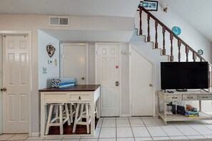 A compact living space with a kitchen area, a dining table with chairs, a television on a stand, and a staircase leading to an upper level. - Leeward Cove B4 - Pool & Bay Views, Dogs Welcome (Corpus Christi)