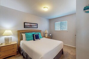 A bedroom with a bed, nightstands, a wall clock, a window with blinds, and a ceiling light. - Leeward Cove B4 - Pool & Bay Views, Dogs Welcome (Corpus Christi)