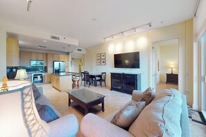 Ein modernes Wohnzimmer mit einer Couch, einem Couchtisch und einem Fernseher. Im Hintergrund befindet sich ein Küchenbereich mit Schränken und einem Kühlschrank.