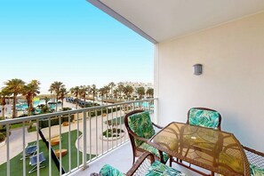 A balcony with wicker furniture and a glass table, overlooking a pool area with palm trees and a clear sky. - Sapphire Condominiums #303 (South Padre Island)