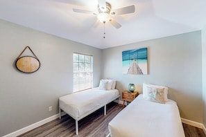 A bedroom with two single beds, a ceiling fan, a window with blinds, and a painting on the wall. - Cora Lee - Private Pool & Putting Green (South Padre Island)