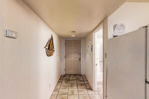 A narrow hallway with a door at the end, a refrigerator on the right, and a wall-mounted light fixture. - Gulfview I #201 - Pool & Hot Tub, Near the Beach (South Padre Island)