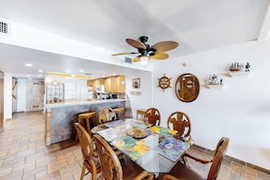 A dining area with a glass-top table, wooden chairs, a ceiling fan, and a kitchen in the background. - Suntide II Condominiums #509 (South Padre Island)