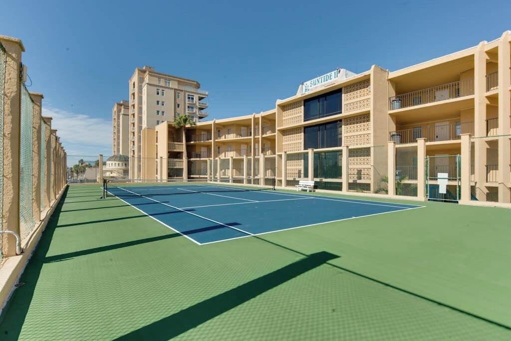 A tennis court with a net, surrounded by buildings with balconies and a sign that reads 'SUNTIDE II'.