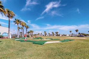 A golf course with a clear sky, palm trees, and a building in the background. - Long Island Village 347 Sand Dollar (Port Isabel)