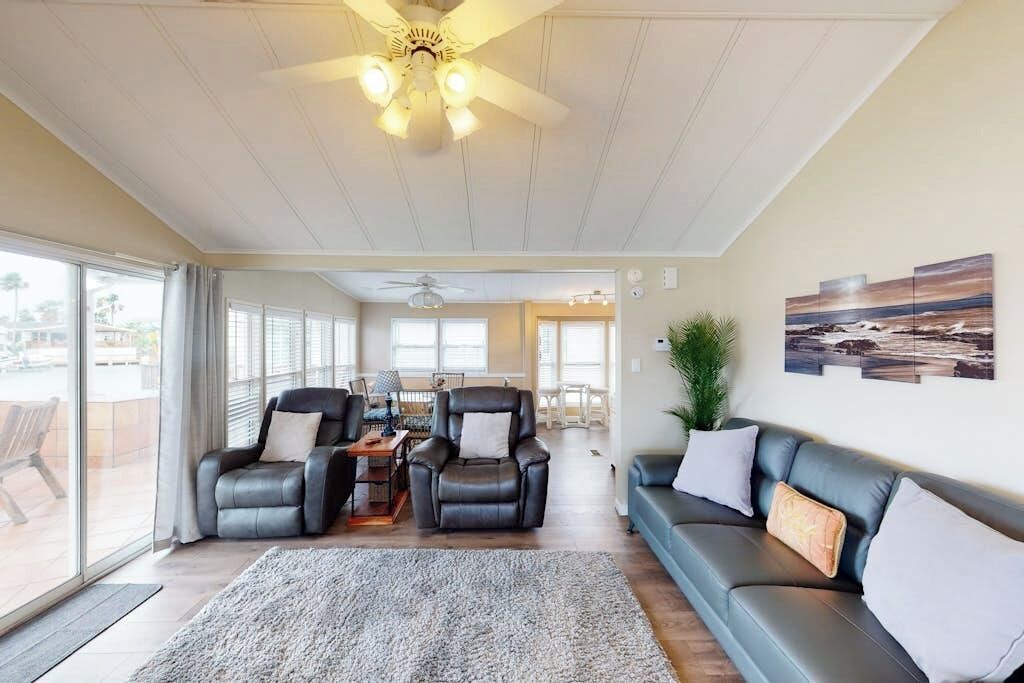 A spacious living room with a large grey rug, two leather sofas, a dining table with chairs, and a ceiling fan.