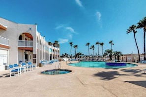 A hotel with a pool area, including a small circular pool and a larger rectangular pool, surrounded by lounge chairs and palm trees. - Long Island Village 347 Sand Dollar (Port Isabel)