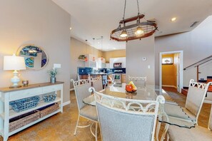 A dining area with a round glass table, white metal chairs, a hanging light fixture, and a console table with decorative items. - Harbor Island Beach House #1533 (Port Isabel)