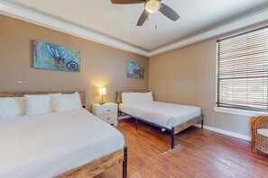 Une chambre avec deux lits, un plancher en bois, un ventilateur de plafond, des œuvres d’art accrochées aux murs et une fenêtre avec des stores.